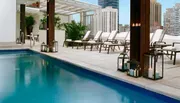 An urban rooftop pool area featuring lounge chairs and decorative lanterns, with a skyline of high-rise buildings in the background.