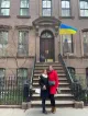 Two smiling women are standing on the steps of a brownstone building under a Ukrainian flag.