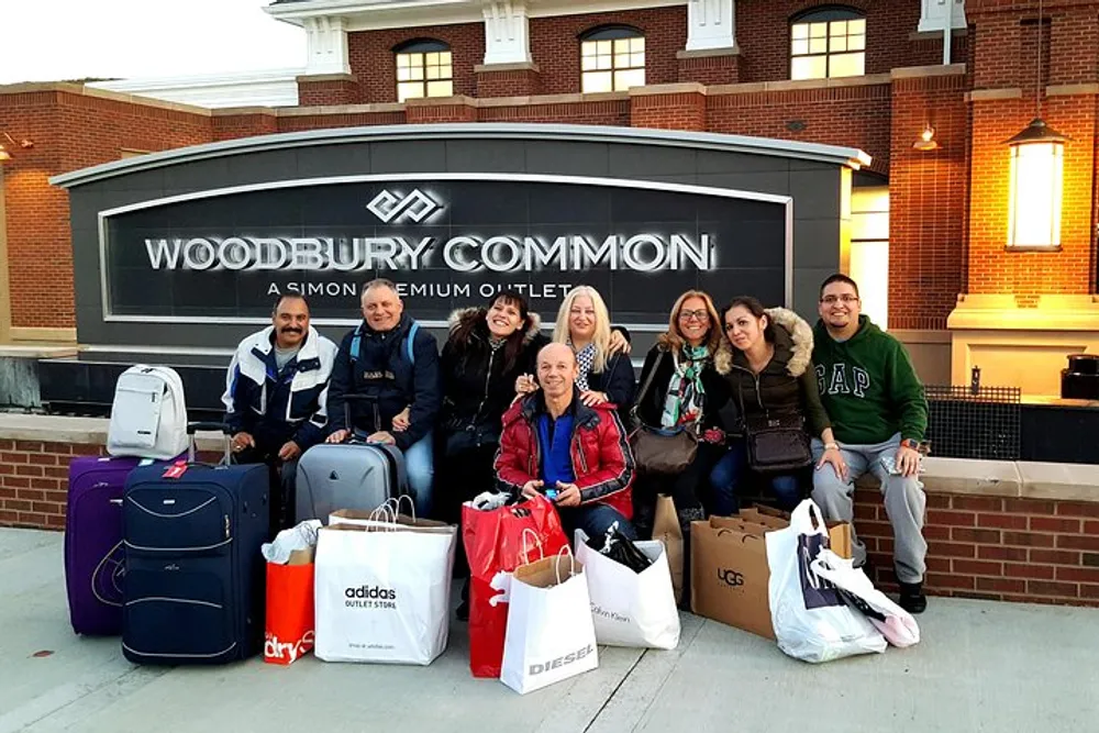 A group of people is posing with shopping bags and luggage in front of the Woodbury Common Premium Outlet sign seemingly happy after a shopping spree