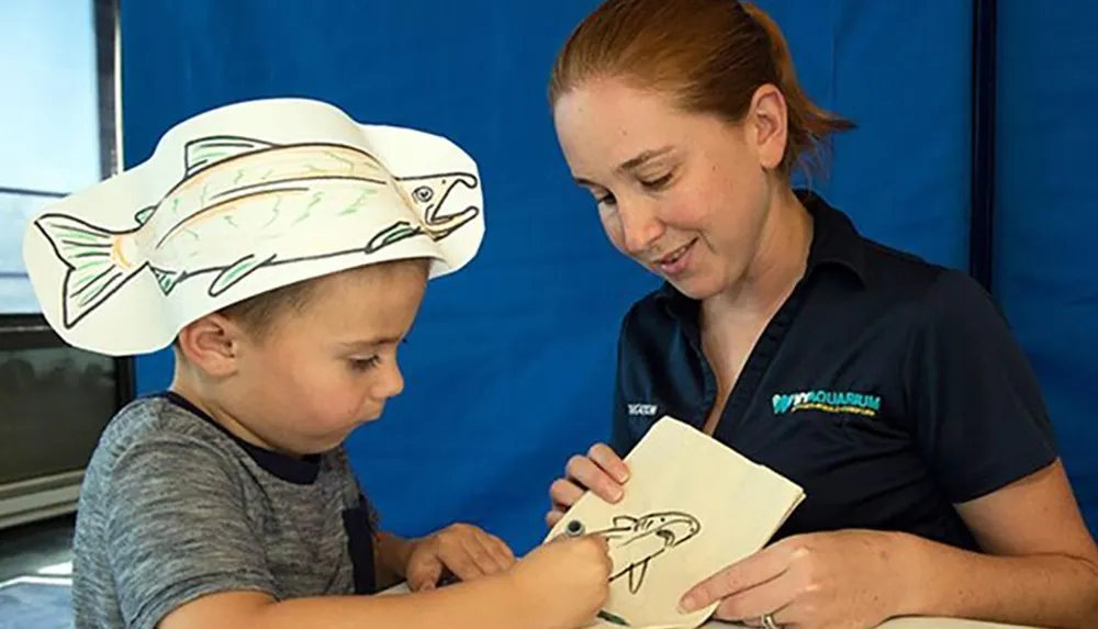 A child and an adult are engaged in an educational activity with the child wearing a fish-shaped hat and both looking at a drawing of a fish