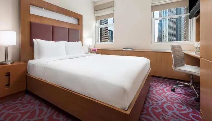 The image shows a neatly arranged hotel room with a large bed modern furnishings and a city view from the window