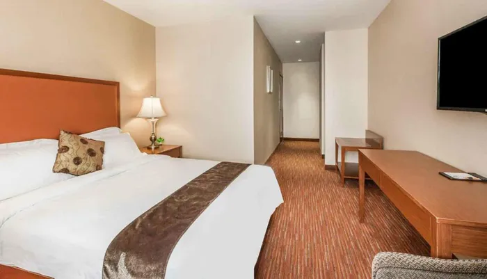 The image is of a neatly arranged hotel room with a large bed side tables a desk a mounted television and a corridor leading to another part of the suite