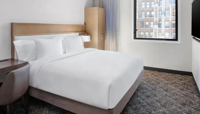 The image depicts a neatly made bed with white linens in a modern hotel room with a view of the adjacent building through the window