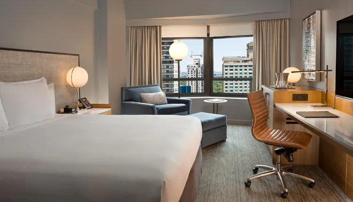 The image shows a modern and neatly arranged hotel room with a large window offering a view of urban buildings