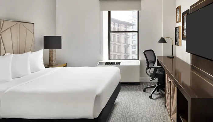 The image shows a modern hotel room with a neatly made bed a work desk with a chair a flat-screen TV on the wall and a view of the city buildings outside the window