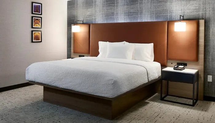 The image shows a modern hotel room with a large bed framed artwork on the wall and a sleek bedside table with a lamp and phone