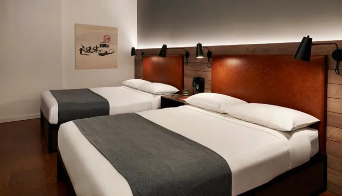 The image shows a modern and neatly arranged twin bedroom with two beds each with white linens and dark grey blankets flanked by wall-mounted lamps against a backdrop of a leather padded headboard and a framed artwork on the wall