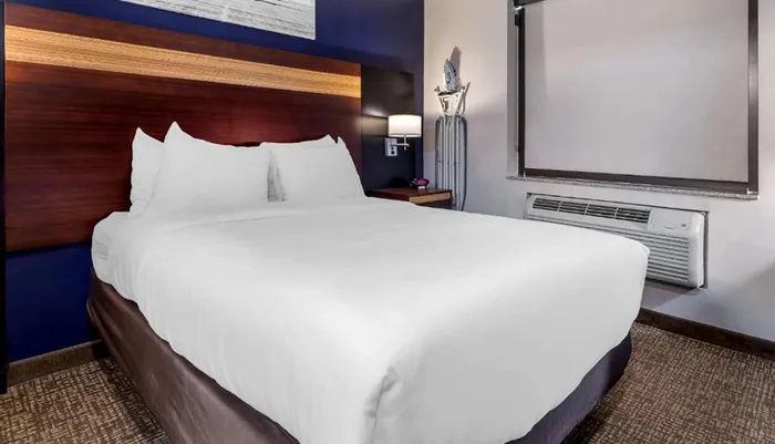 The image shows a neatly made bed in a modern hotel room with simplistic decor and a wall-mounted air conditioning unit