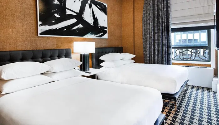 The image shows a modern hotel room with two queen-sized beds an abstract black and white painting above the headboard a large window and a patterned carpet