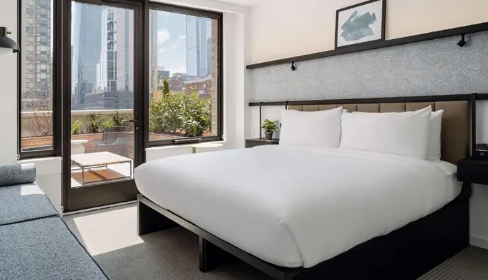 The image shows a modern bedroom with a large bed sleek furniture a balcony and a view of urban high-rises under a clear sky