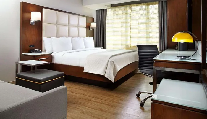 The image shows a modern hotel room with a neatly made bed a desk with a chair and a window with striped curtains