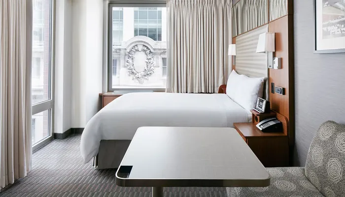 The image shows a well-lit modern hotel room with a large bed elegant decor and a window overlooking architectural details of an adjacent building