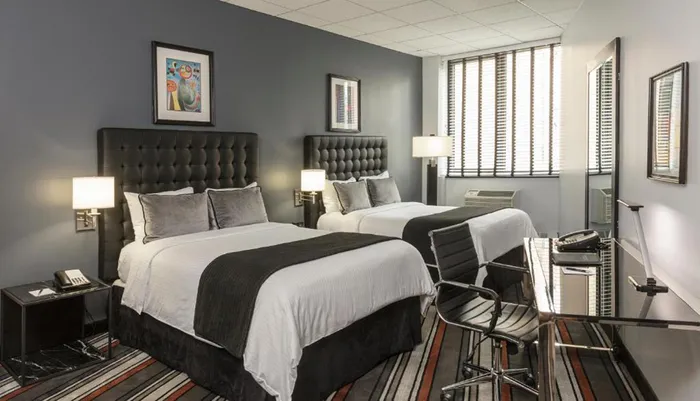 The image shows a well-lit modern hotel room with two neatly made beds a desk with a chair striped carpet dark grey walls and framed artwork