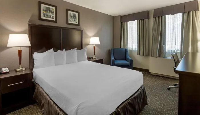 This is an image of a neatly arranged hotel room featuring a large bed with white linens a working desk with a chair an armchair and framed pictures above the bed