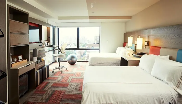 The image shows a modern hotel room with two beds a work desk a balance ball chair and a view of the city through a large window