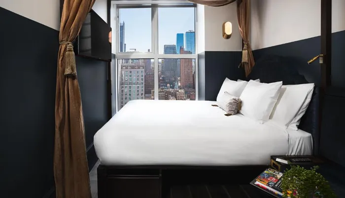 A neatly made bed with white linens is set against a dark blue headboard with a large window showcasing an urban skyline