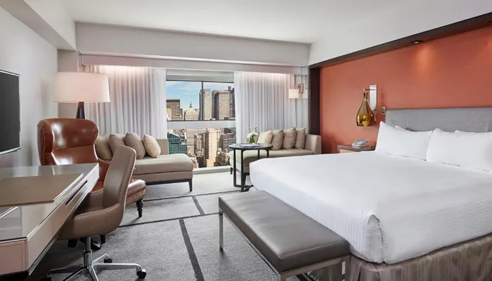The image shows a modern hotel room with a large bed a workspace a sitting area and a window offering a view of a city skyline
