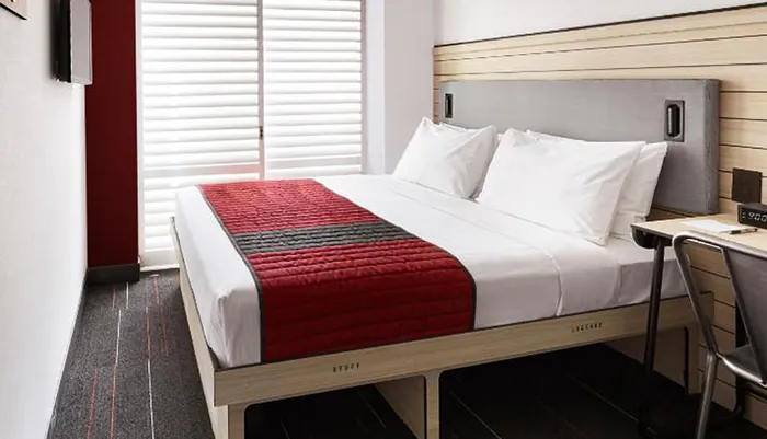 The image shows a neatly made bed with white linens and a red bed runner in a modern hotel room with a work desk and closed blinds on the window