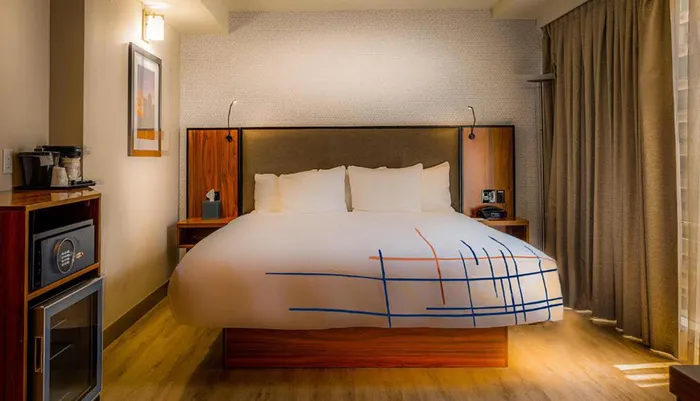 The image shows a neatly arranged hotel room with modern furnishings and a large bed with a distinctive blue and orange line pattern on the duvet