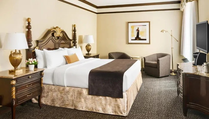 The image shows a well-appointed hotel room with classic-style furniture a plush bed elegant lamps and abstract art on the wall