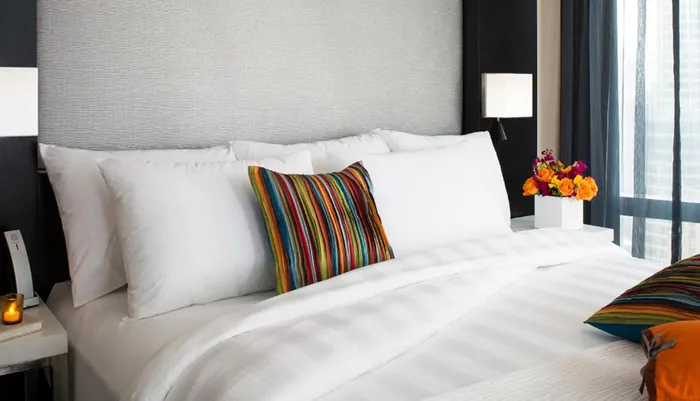The image showcases a neatly made bed with white bedding and colorful accent pillows accompanied by a bedside lamp and a bouquet of flowers within a modern room setting