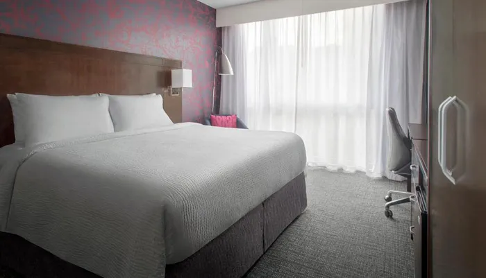 The image shows a neatly arranged modern hotel room with a large bed subtle lighting a patterned accent wall and a desk area with a chair near the window covered by sheer curtains