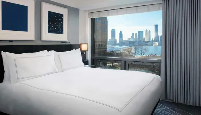 A modern hotel room with a large bed offers a panoramic view of a city skyline through its window