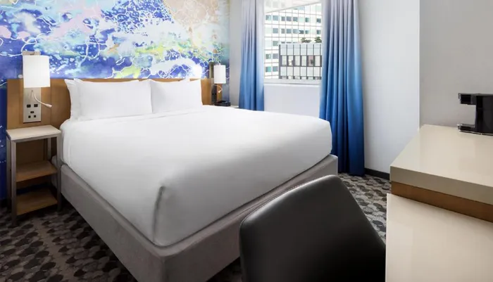 The image shows a modern hotel room with a neatly made bed abstract wall art and a view of an urban exterior through the window