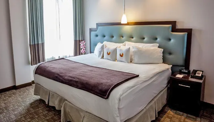 This image shows a neatly arranged hotel room with a large bed crisp white bedding accentuated with a burgundy throw and decorative pillows and minimalist decor