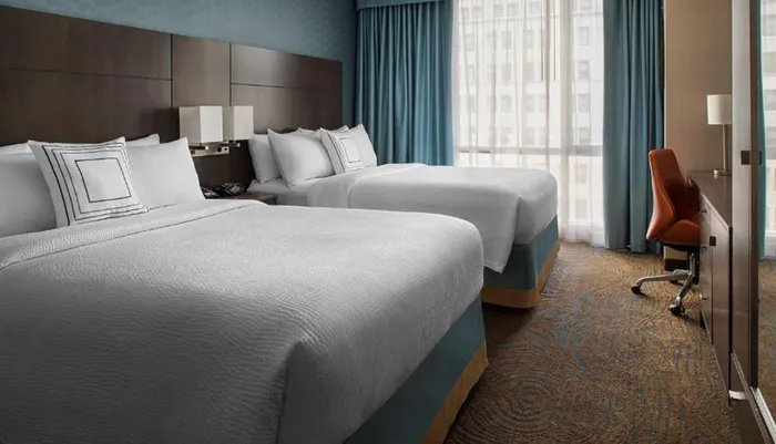 The image shows a modern hotel room with two neatly made queen-sized beds accentuated by a patterned carpet sheer curtains with a view of adjacent buildings and a workspace featuring an ergonomic chair