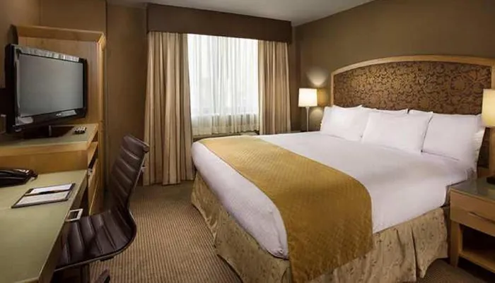 The image shows a well-appointed hotel room with a neatly made bed a flat-screen TV a work desk with a chair and curtains drawn back to allow light into the room
