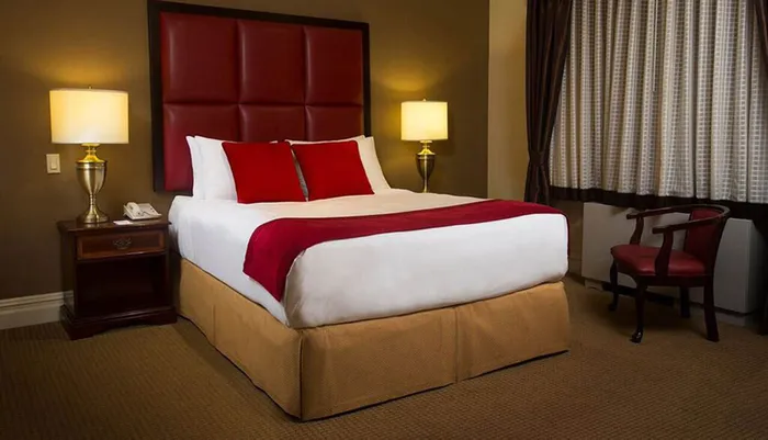 The image shows a neatly arranged hotel room with a large bed featuring white linens and red accents flanked by matching table lamps and a desk with a chair near a curtained window