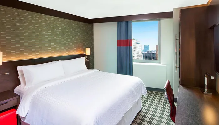 This image shows a modern hotel room with a large bed stylish decor and a window offering a view of an urban skyline
