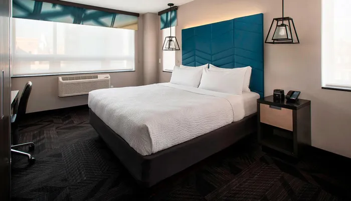 A modern hotel room featuring a large bed with a blue headboard clean white bedding unique hanging lamps and a window with a blurred city view