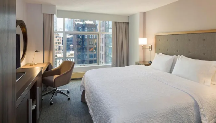 The image shows a modern hotel room with a large bed a work desk with a chair and a view of urban buildings through the window