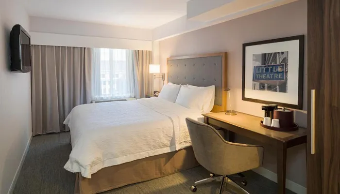 The image shows a neatly arranged modern hotel room with a large bed a desk and chair a mounted television and a framed picture on the wall