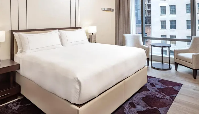 The image shows a neatly made bed in a modern and well-lit hotel room featuring a seating area by the window