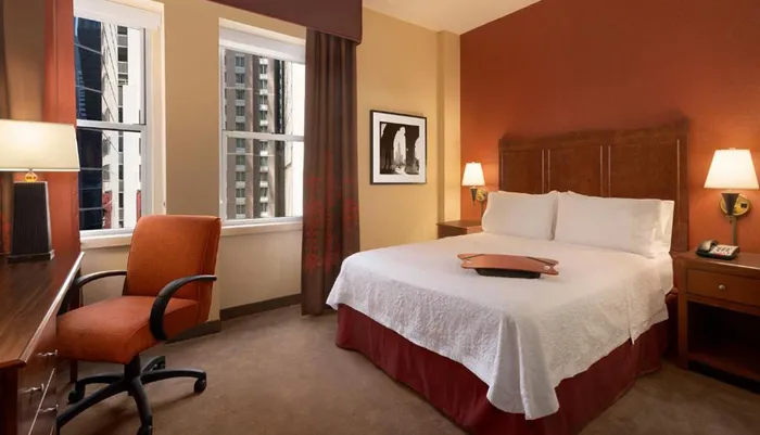 The image shows a neatly arranged hotel room with a large bed a desk with an orange chair two windows with a city view and warm lighting creating a cozy atmosphere