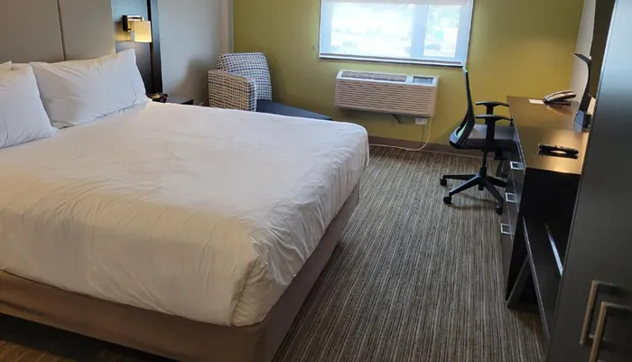 The image shows a neatly arranged hotel room with a large bed a desk with an office chair and a greenish-yellow wall