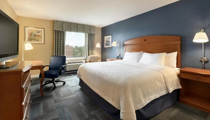 The image shows a neatly arranged hotel room with a king-sized bed a working desk with a chair a flat-screen TV and a window with a view of the outside