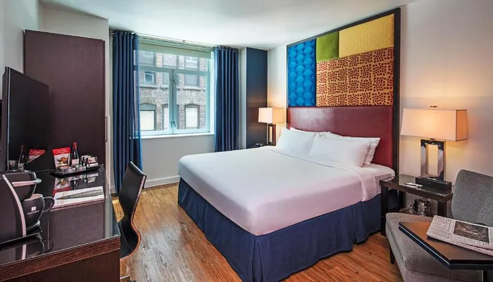 A modern hotel room featuring a large bed with a colorful headboard a working desk a seating area and a window with a view of an adjacent building
