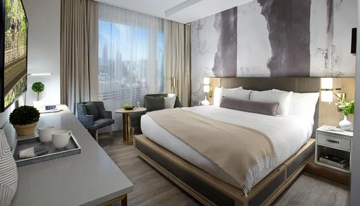 The image shows a modern and well-appointed hotel room with a large bed seating area and a view of the city skyline through the window