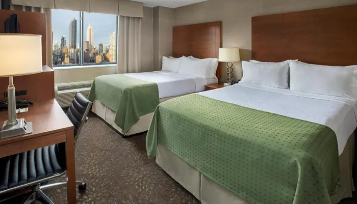 The image shows a neatly arranged hotel room with twin beds contemporary furnishings and a large window offering a city skyline view