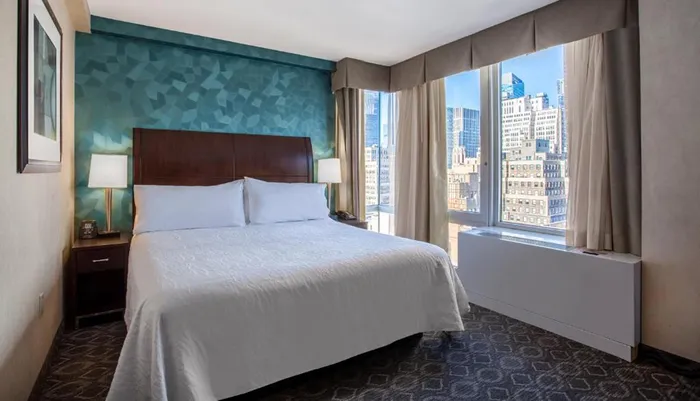 A modernly furnished hotel room with a large bed and a view of a city skyline through the window