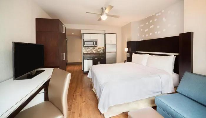 The image shows a modern hotel room with a bed kitchenette and a seating area