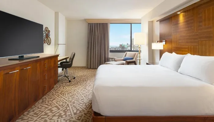 This image shows a neatly organized hotel room with a large bed a desk and chair a television and a window offering a view of the buildings outside