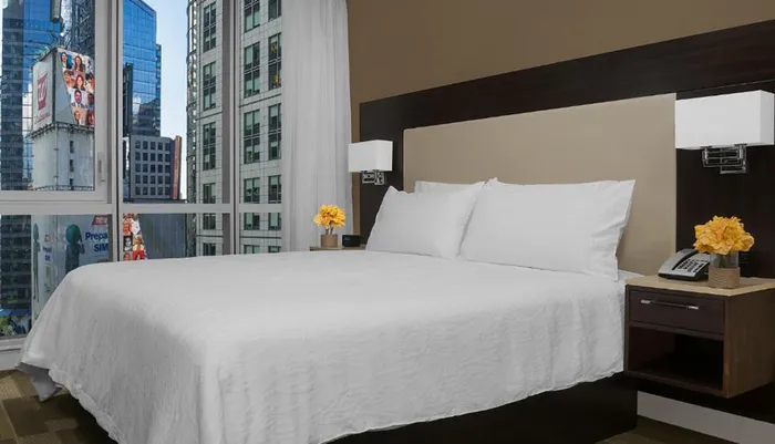 A neatly made bed with white linens is centered in a modern hotel room with a view of a cityscape through the window