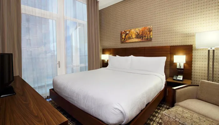 A neatly arranged hotel room with a large bed patterned wallpaper a painting of autumn trees and modern amenities bathed in natural light from a window