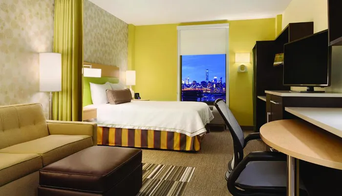 This image features a modern hotel room with a cozy bed a seating area a workspace and a large window that offers a stunning night view of a city skyline