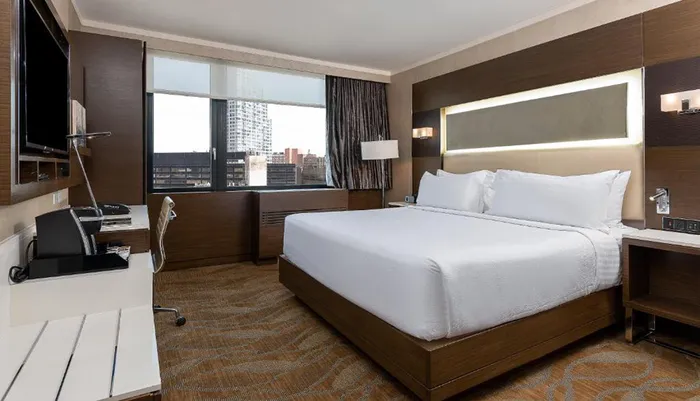 The image shows a modern hotel room with a large bed a work desk a flat-screen TV and a view of urban buildings outside the window